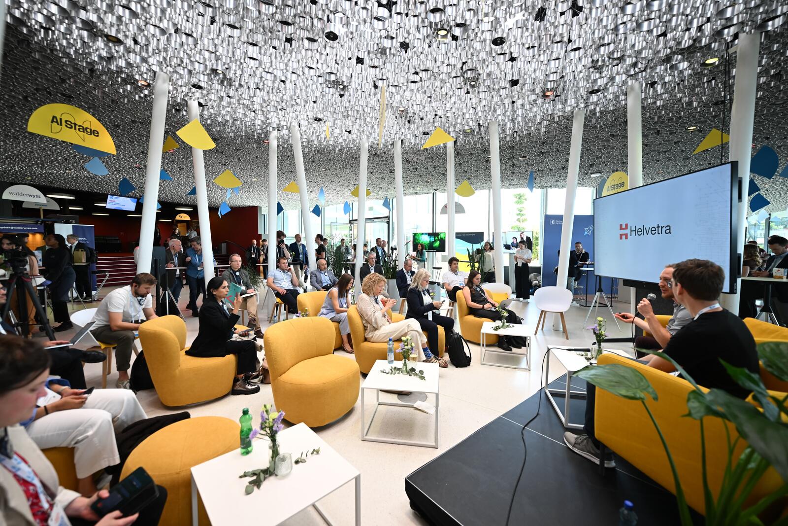 The Helvetra logo on a large screen at the AI Stage of Swiss Startup Days 2026, Kursaal Bern. Audience seated on yellow lounge chairs; Alex (right of frame) speaks into a microphone next to Oleg Lavrovsky.
